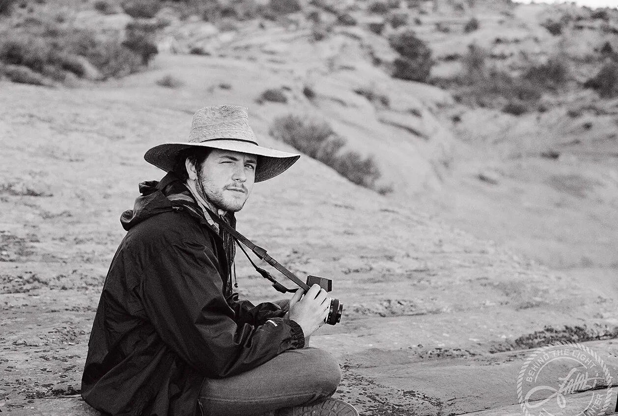 Black-and-white photo of John Matthew Gillen in a wide-brimmed hat, holding a film camera in the desert